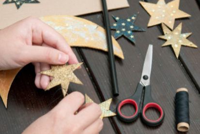 Table top filled with crafting supplies and someone is cutting out golden glitter star shapes.